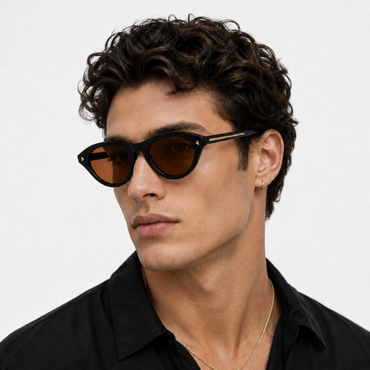 A man with curly dark hair wearing black cat-eye sunglasses with amber lenses and a black shirt, photographed against a light gray studio background.